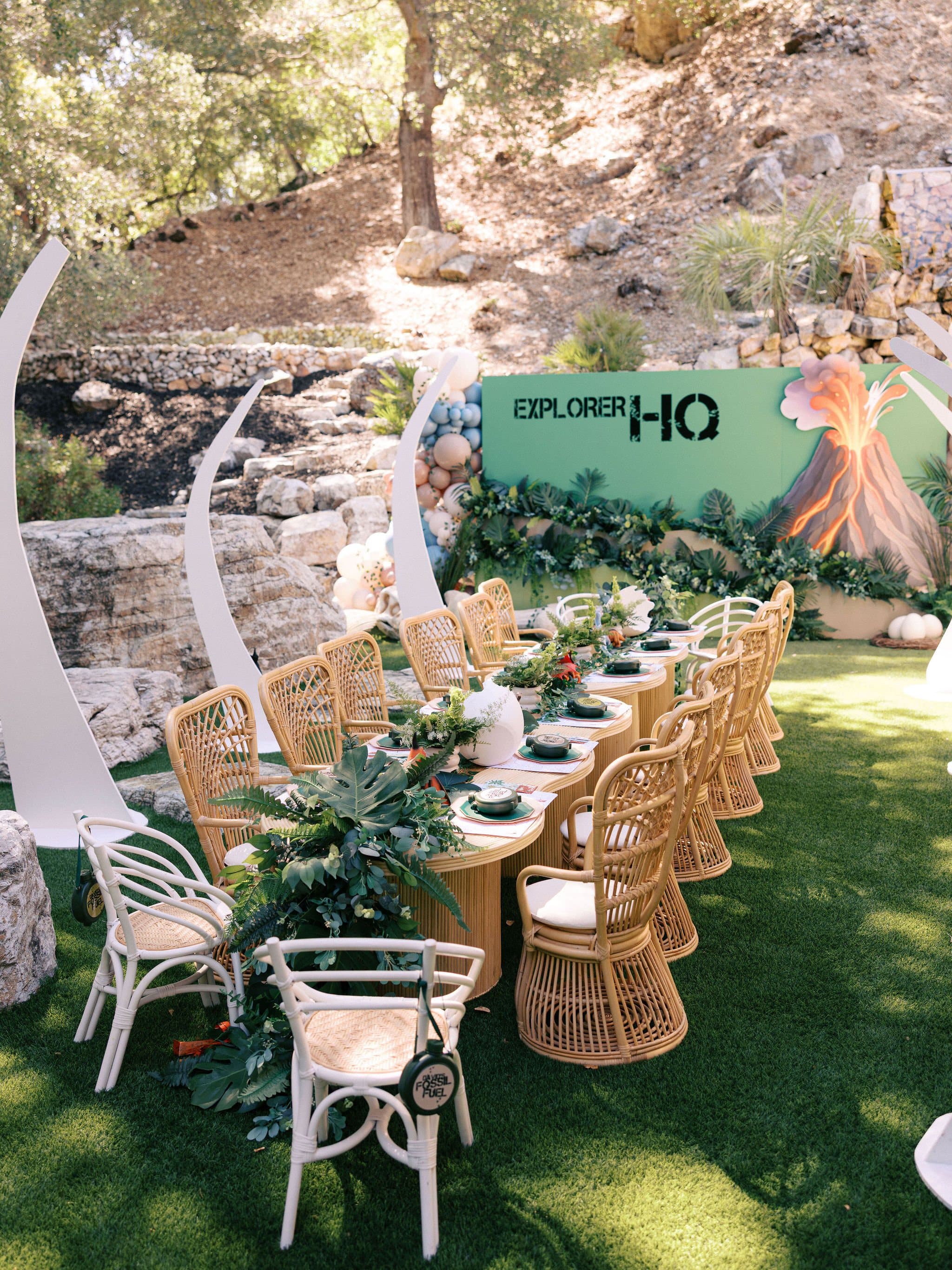 Long kids table with rattan chairs under the trees at a dinosaur party
